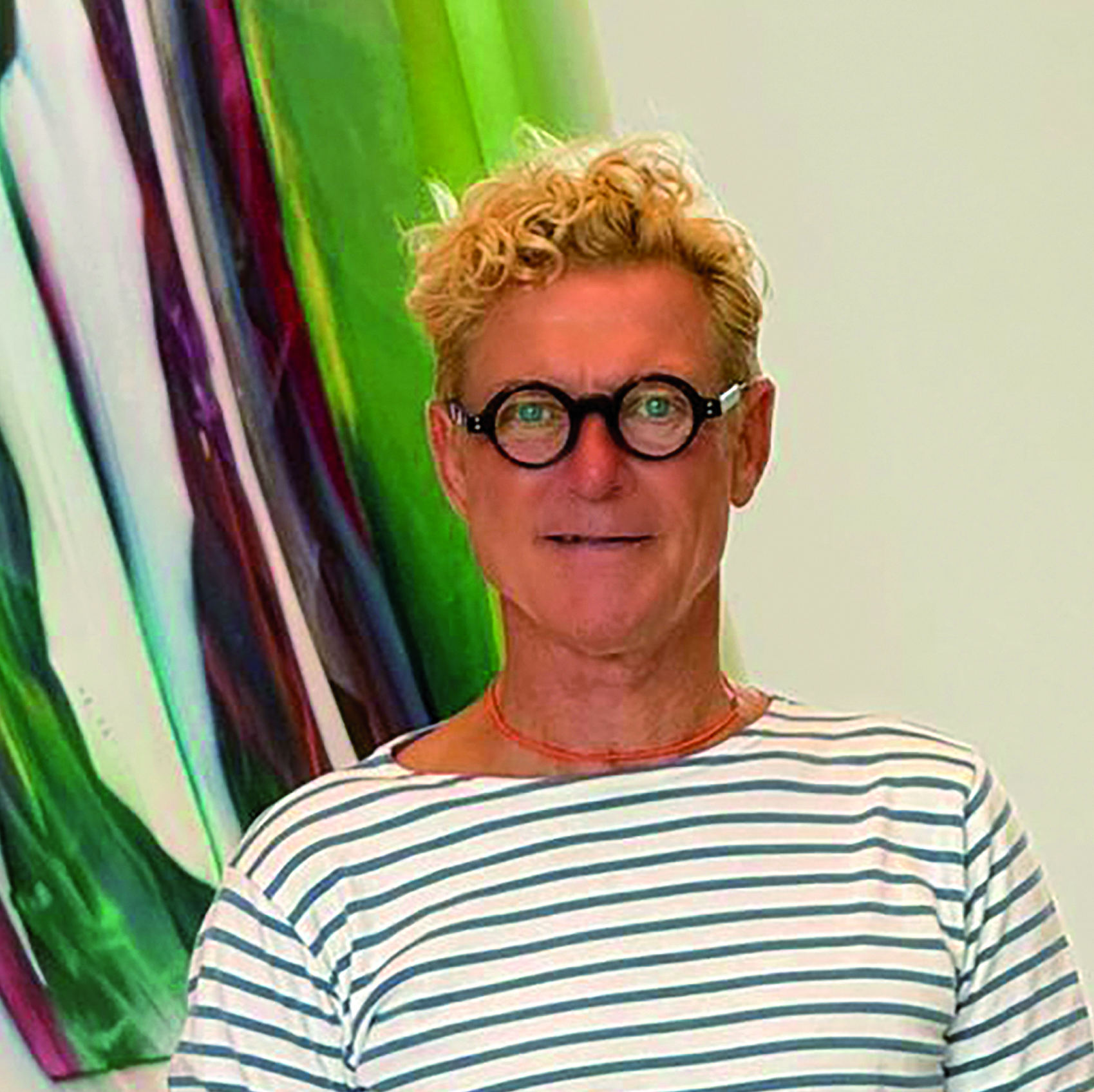 Portrait of Markus Böhler in a Breton-striped shirt and round glasses, a colourful painting behind him